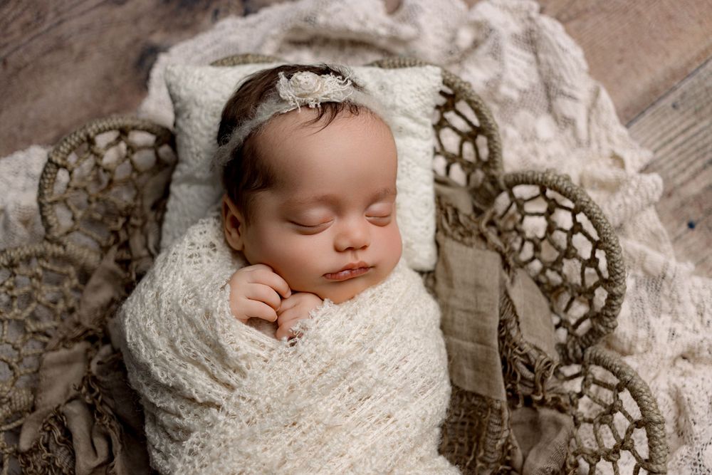 Sleeping newborn wrapped in white lace swaddle resting in textured neutral basket with delicate lace headband.