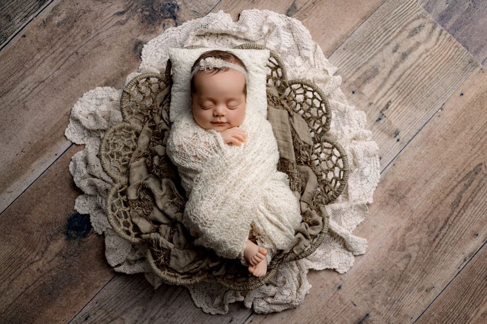 Newborn baby wrapped in white swaddle lying on circular lace doily on wooden floor.