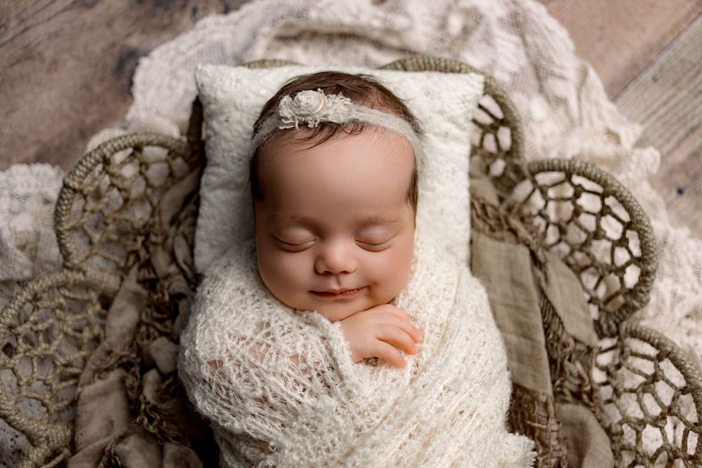 A sleeping newborn wrapped in soft white knit swaddle lies peacefully on lace textured fabric.
