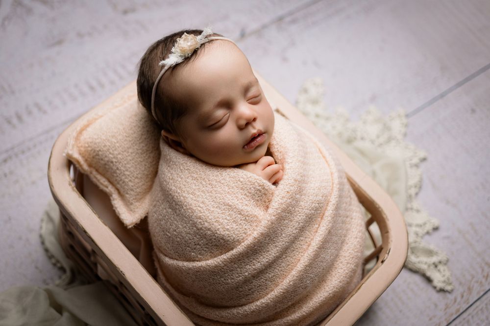 Sleeping newborn baby wrapped in beige blanket lying in wooden basket wearing delicate headband.
