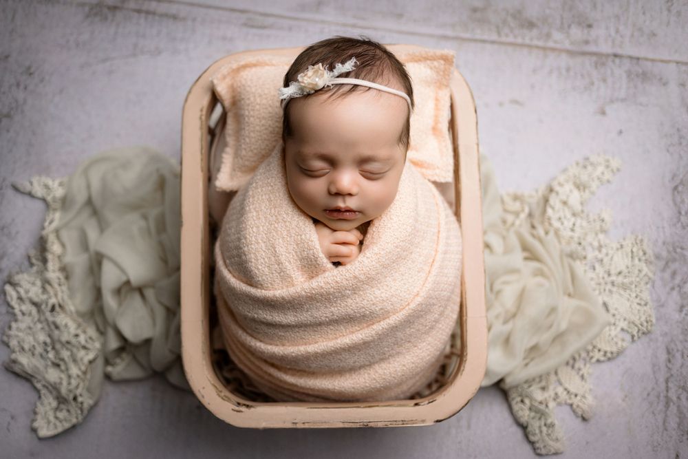 A peaceful sleeping newborn swaddled in soft peach fabric lies in a wooden basket surrounded by delicate lace fabric.