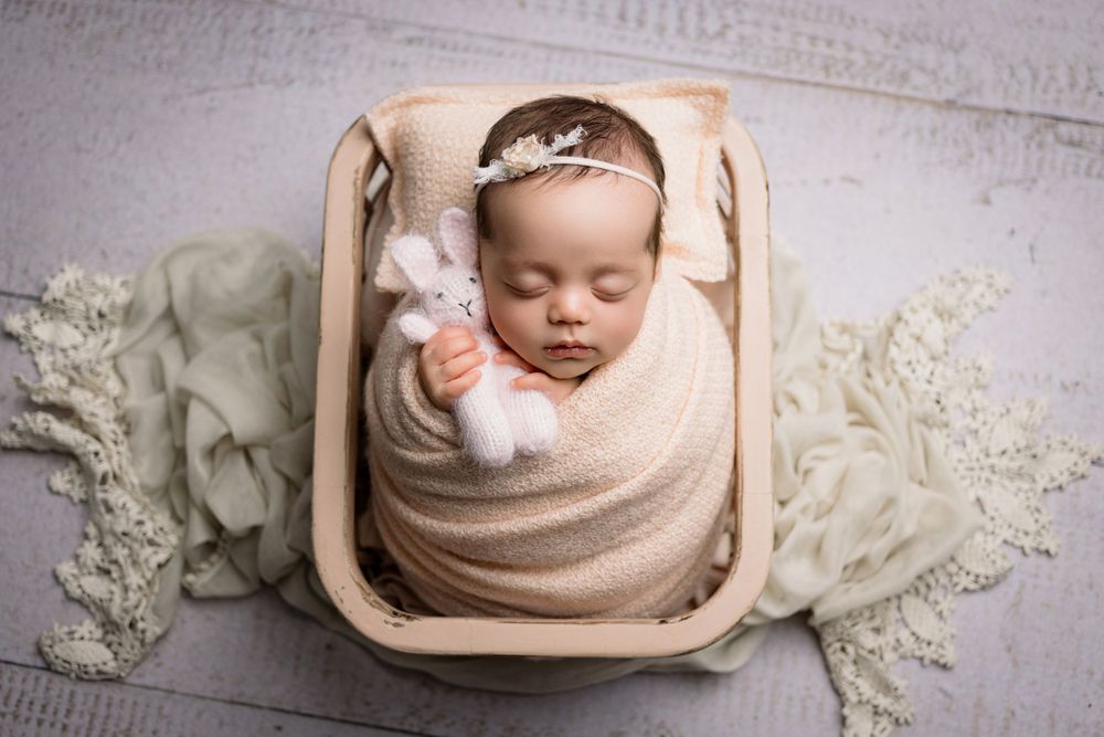 A peaceful sleeping newborn wrapped in beige fabric lies in a wooden bowl surrounded by delicate white flowers and lace.