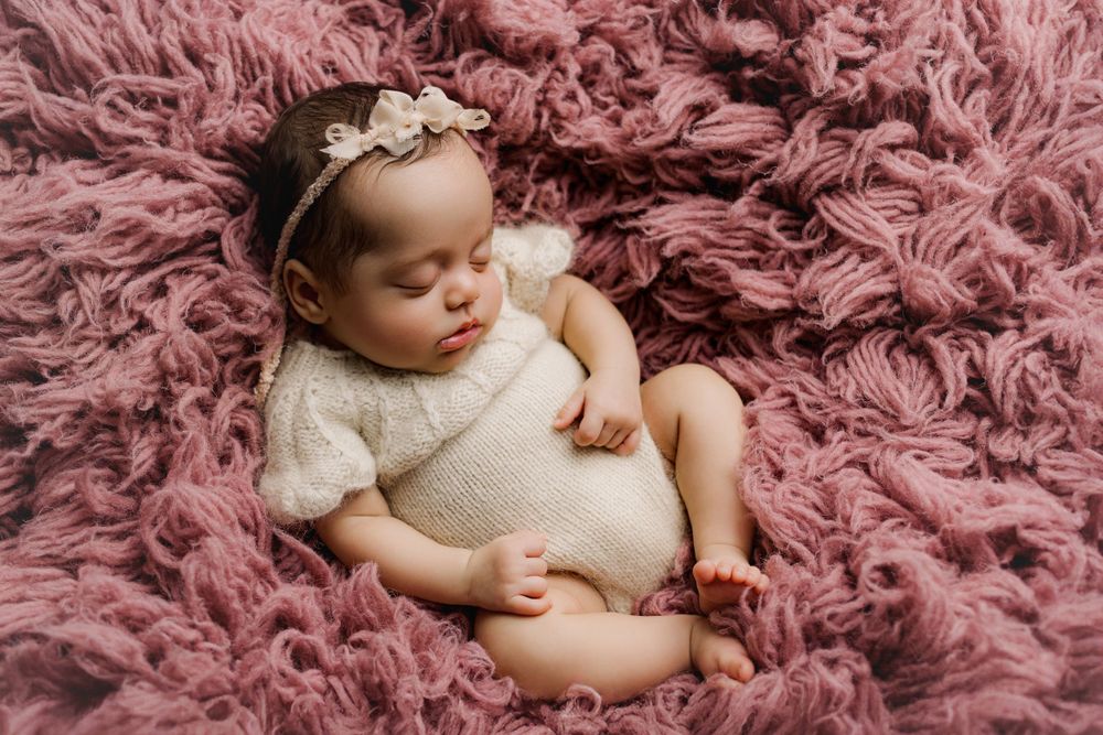 Sleeping newborn wearing white lace outfit and floral headband rests peacefully on pink fluffy blanket.