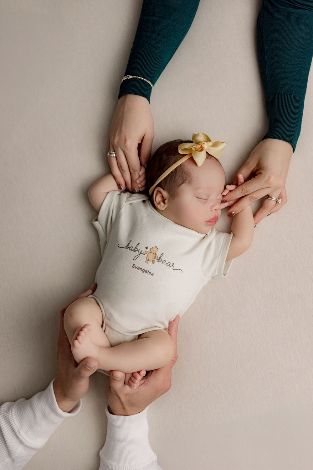 A sleeping baby in white onesie with gold bow headband being gently cradled by hands wearing dark green sleeves.