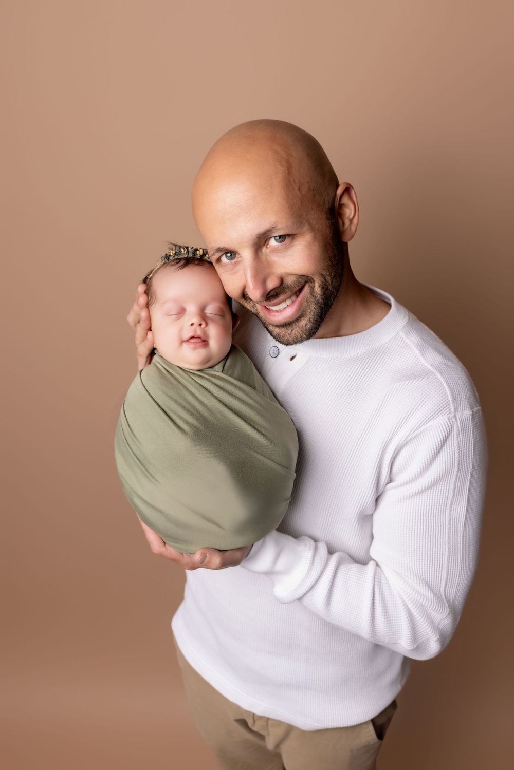 A heartwarming portrait against a brown backdrop showing a person in a white sweater tenderly holding a wrapped newborn.