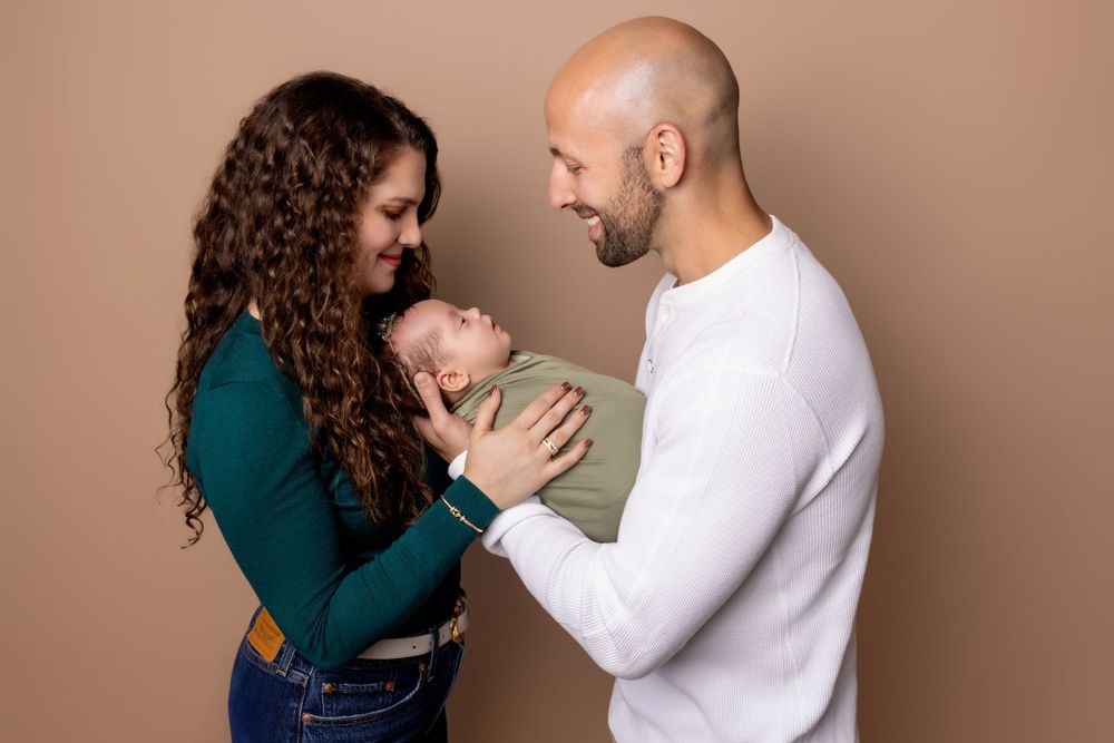 Parents in white and green tops tenderly holding their newborn against brown background.