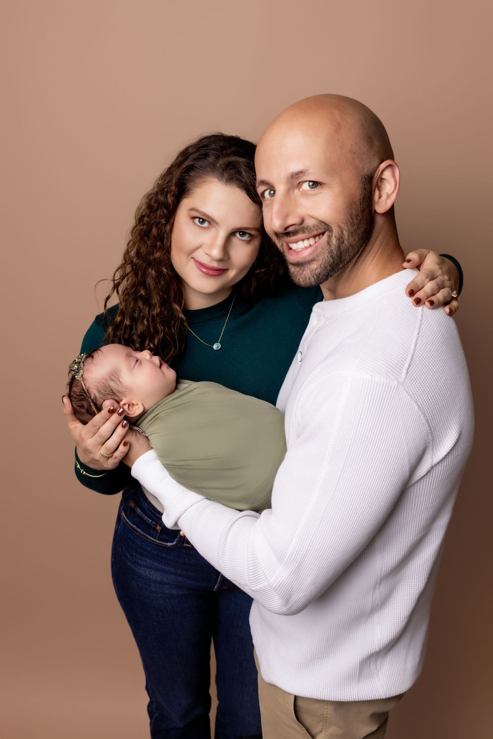 A couple embraces while holding a newborn baby wrapped in sage green cloth against a brown studio backdrop.