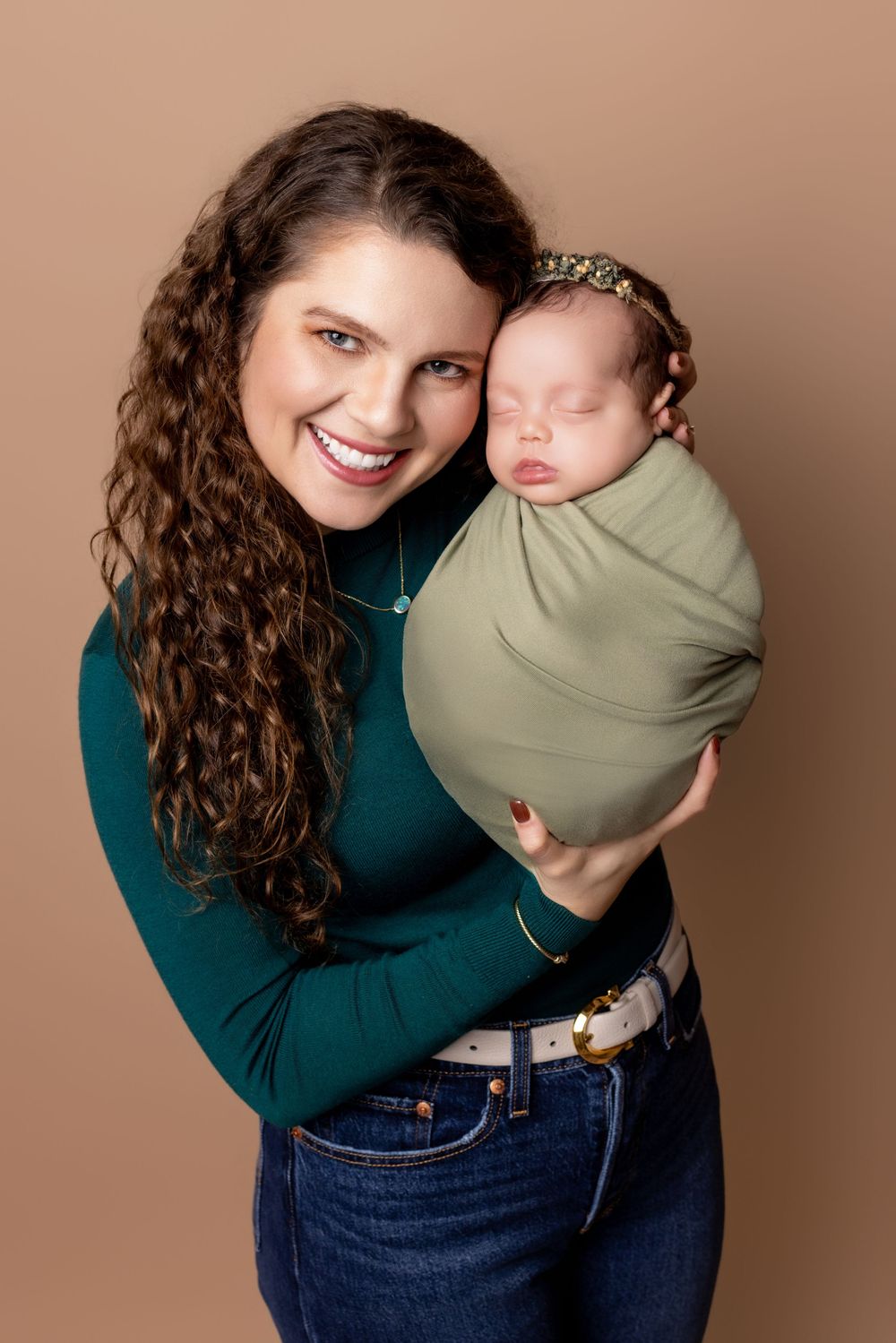 Studio portrait showing a newborn baby swaddled in sage green fabric with a tiny crown against a warm brown background.