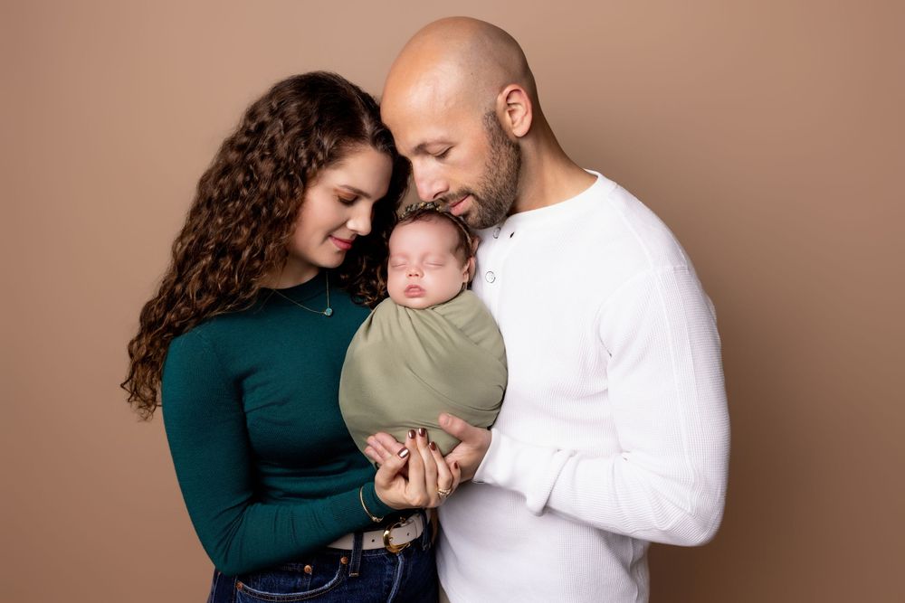 Family portrait with couple embracing their newborn baby against brown backdrop.