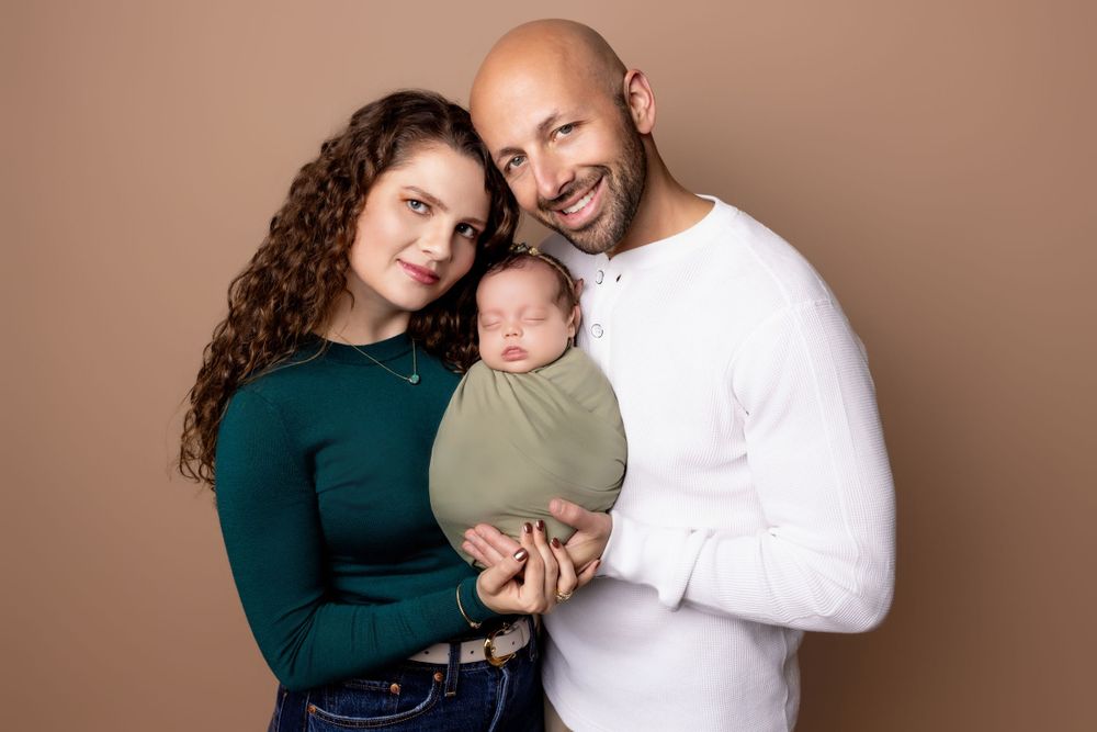 Family wearing green and white clothing holds newborn baby wrapped in sage green swaddle against brown backdrop.