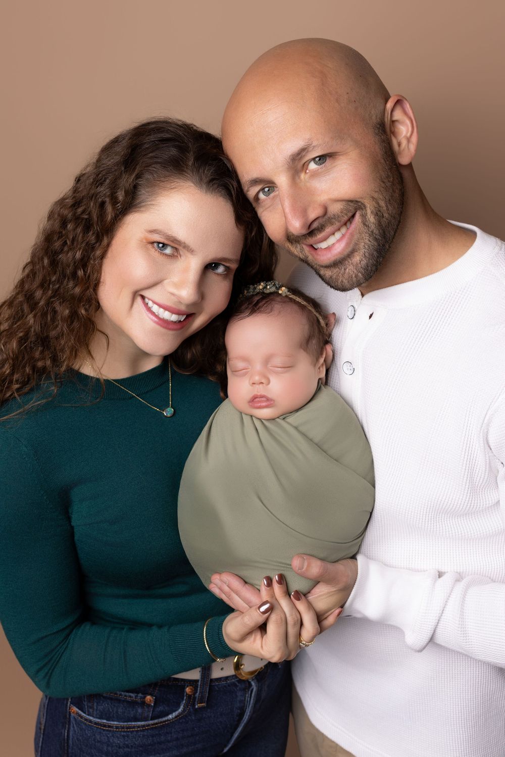 A couple in casual clothing holds their sleeping newborn baby against a neutral brown studio backdrop.