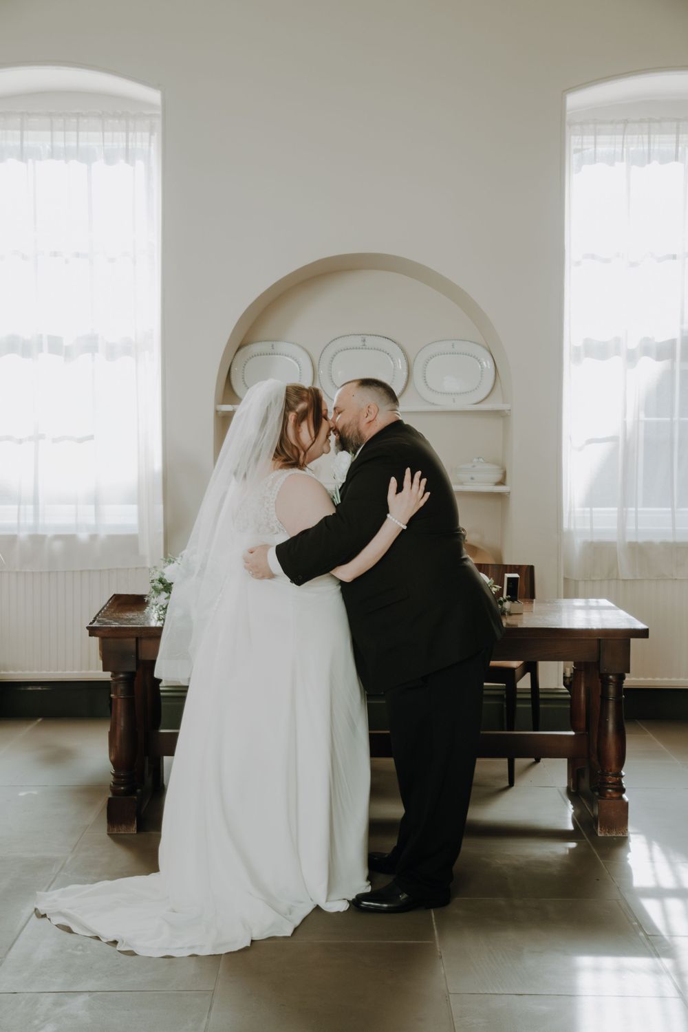 A couple shares an intimate moment in a bright room with white curtains and vintage wooden furniture in the background.