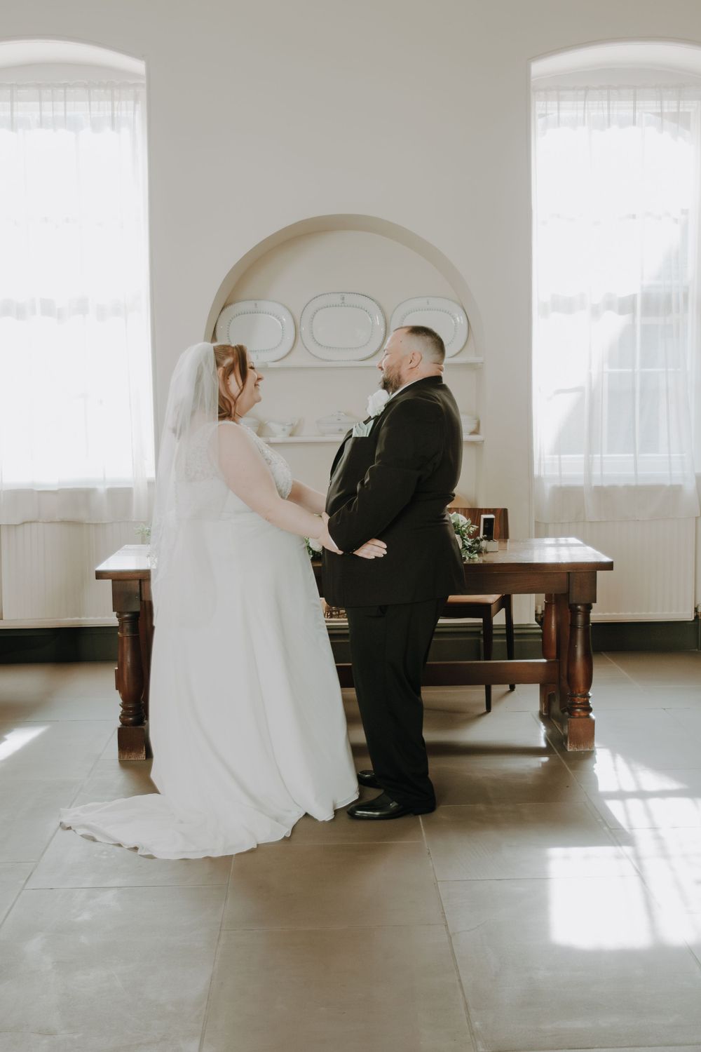 A romantic wedding photo showing a couple standing in an elegant room with arched windows and wooden furniture.
