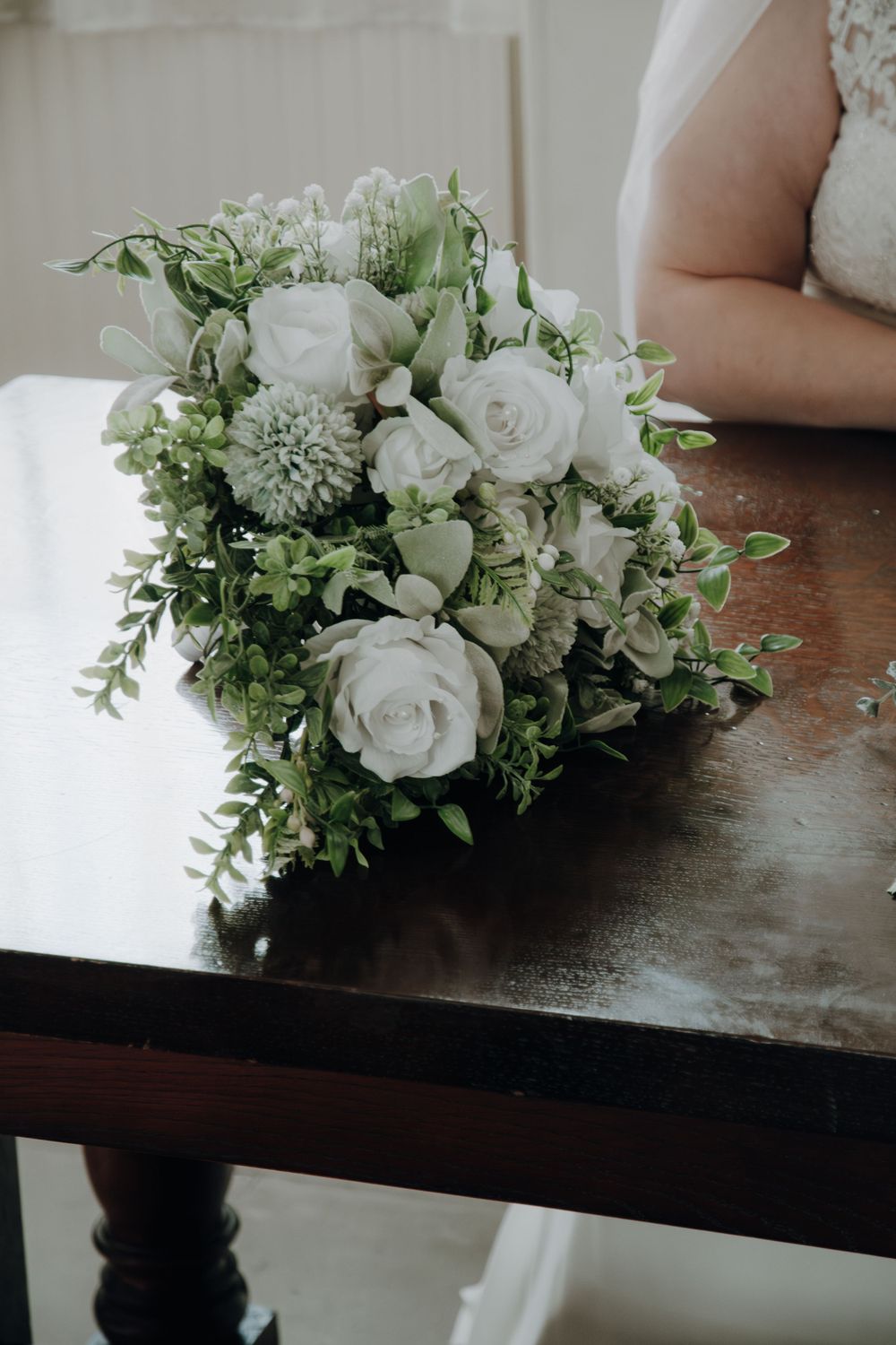 A delicate bridal bouquet with white roses, green foliage and small hydrangeas rests on a dark wooden table surface.
