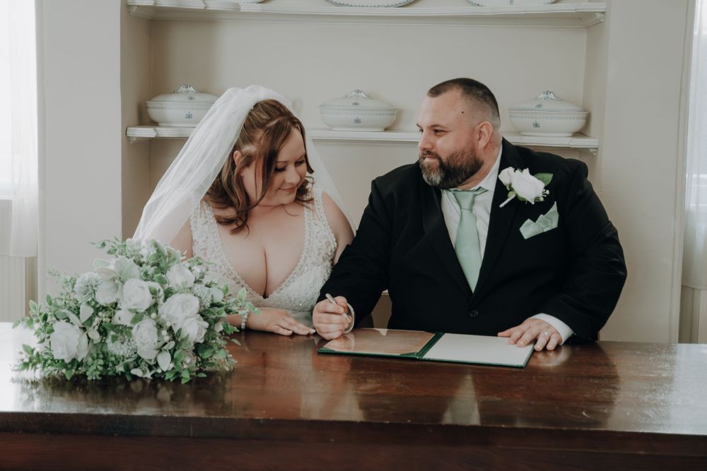 Newlyweds sign their marriage certificate at wooden table decorated with white floral arrangements.