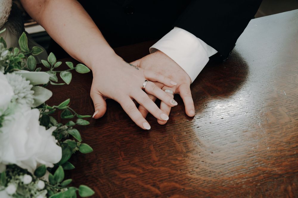 White-clad hands wearing a ring rest on a dark wooden table next to green foliage and white flowers.