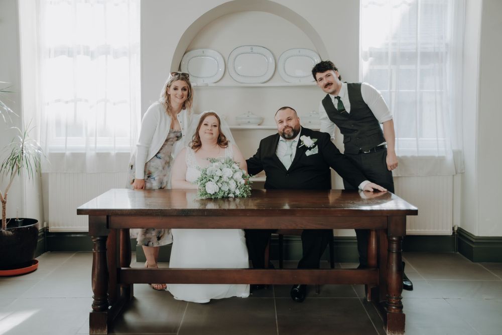 Wedding party poses playfully at a rustic wooden table during a bright indoor celebration.