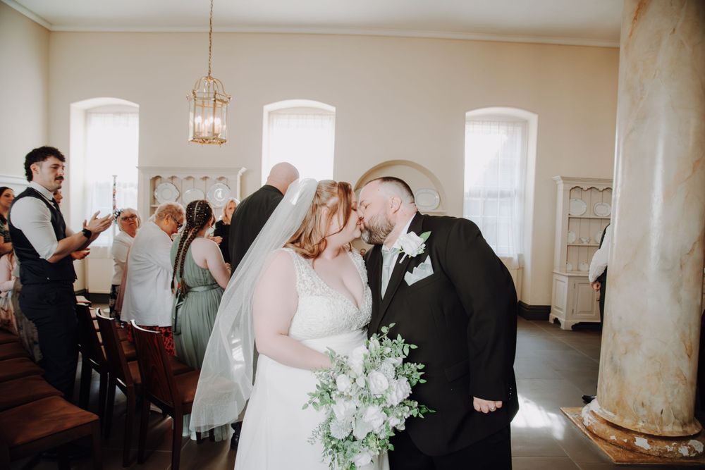 A couple shares a tender moment during their wedding ceremony in an elegant room with arched windows and marble columns.