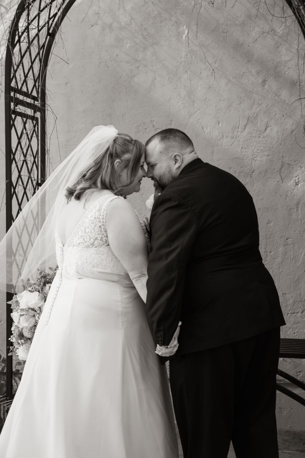 A tender black and white wedding portrait of a couple sharing an intimate moment by a latticed window.