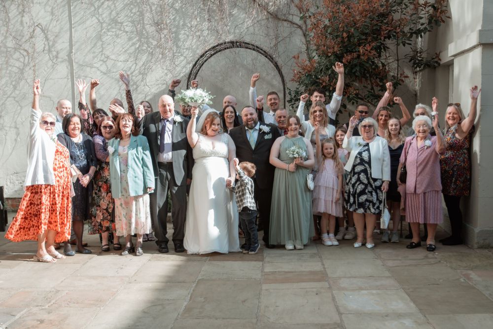 Large wedding party group poses joyfully with raised arms under stone archway at outdoor event celebration.