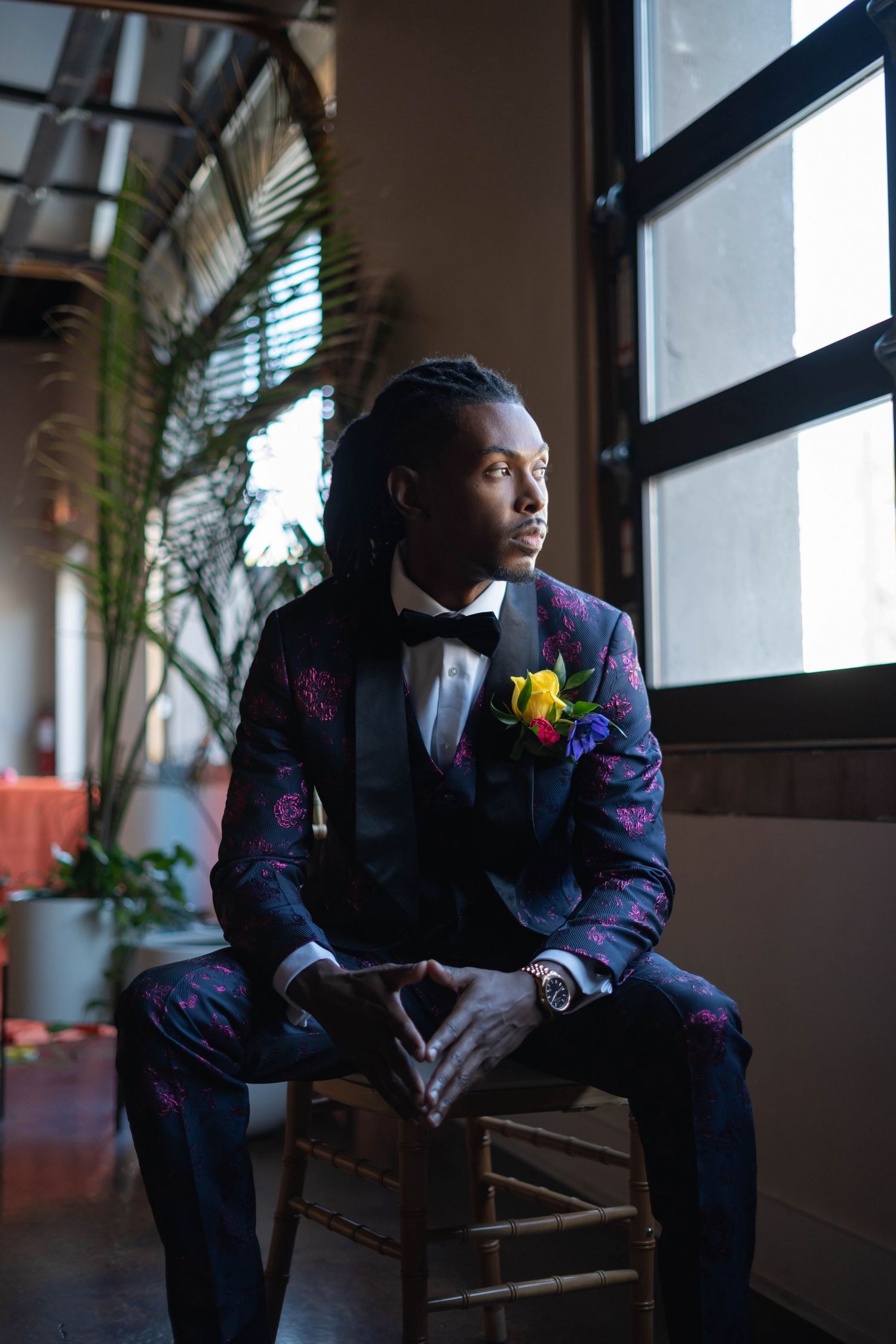 A person in a stylish floral purple suit with bowtie and yellow boutonniere sits thoughtfully by a window.