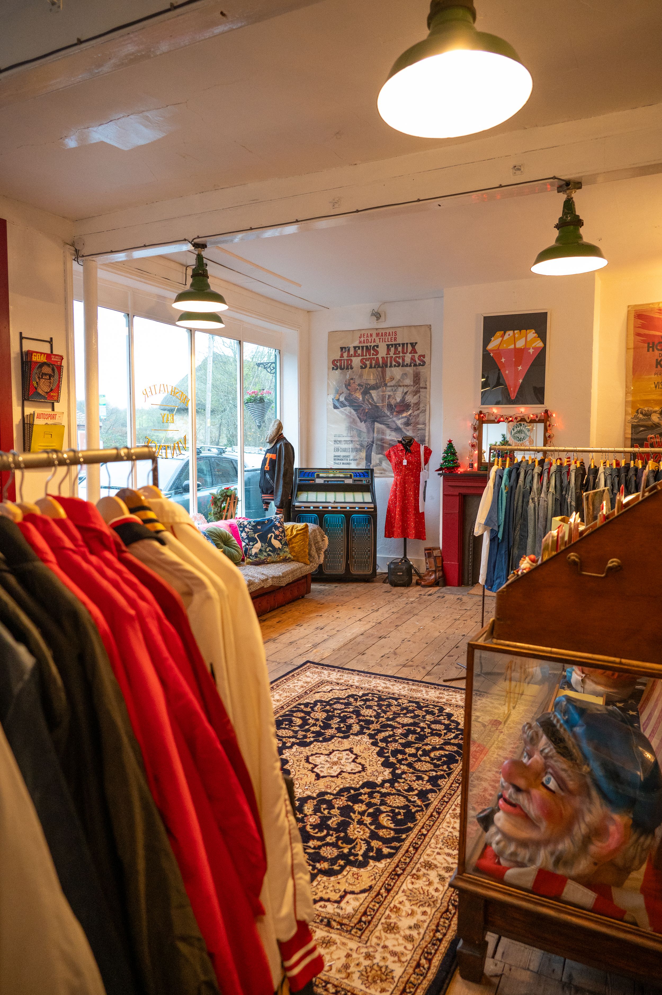 Interior of a vintage clothing store with hanging garments, patterned rugs, and retro pendant lighting creating a cozy atmosphere.