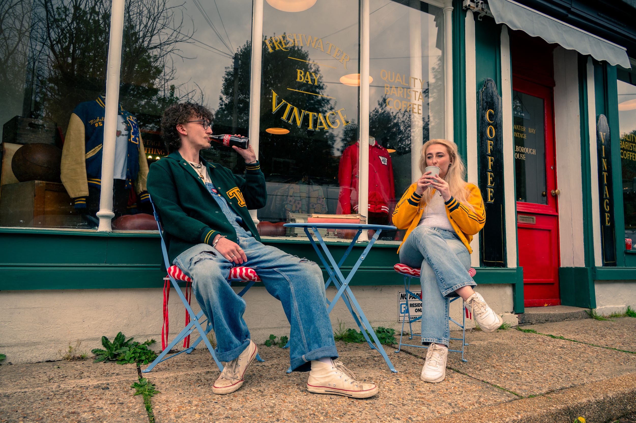 Two friends share a casual meal while sitting at outdoor cafe table near storefront window in casual attire.