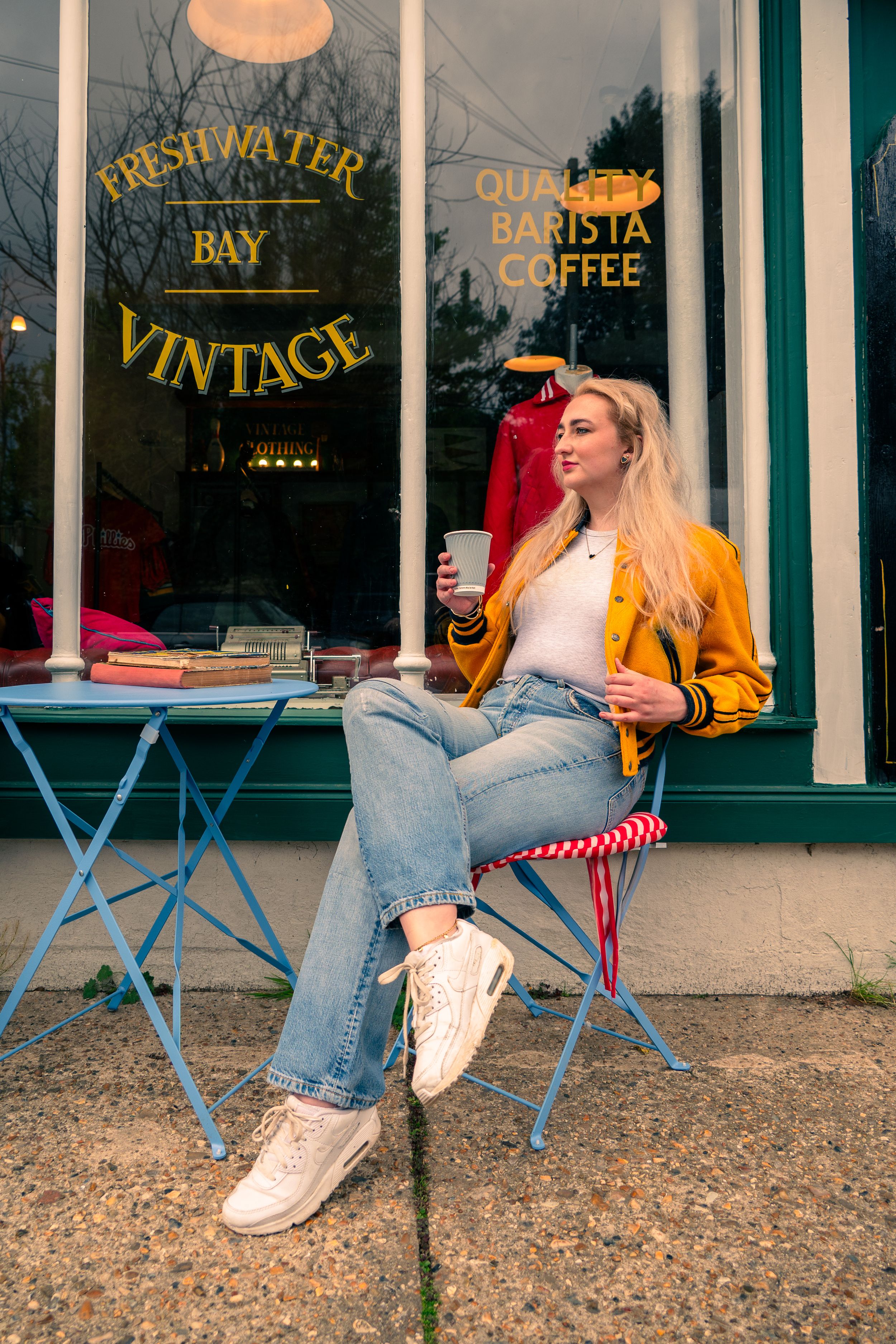 Someone sits outside a vintage coffee shop in a yellow jacket and jeans, enjoying a drink at a blue bistro table.