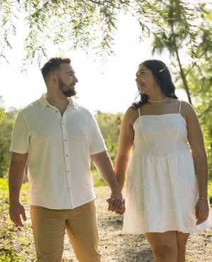 A couple holding hands walk along a sunlit nature path, she in a white sundress and he in a cream button-down shirt.