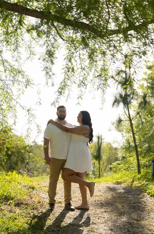 A couple in white outfits share a tender moment while standing on a sunlit nature path under hanging tree branches.