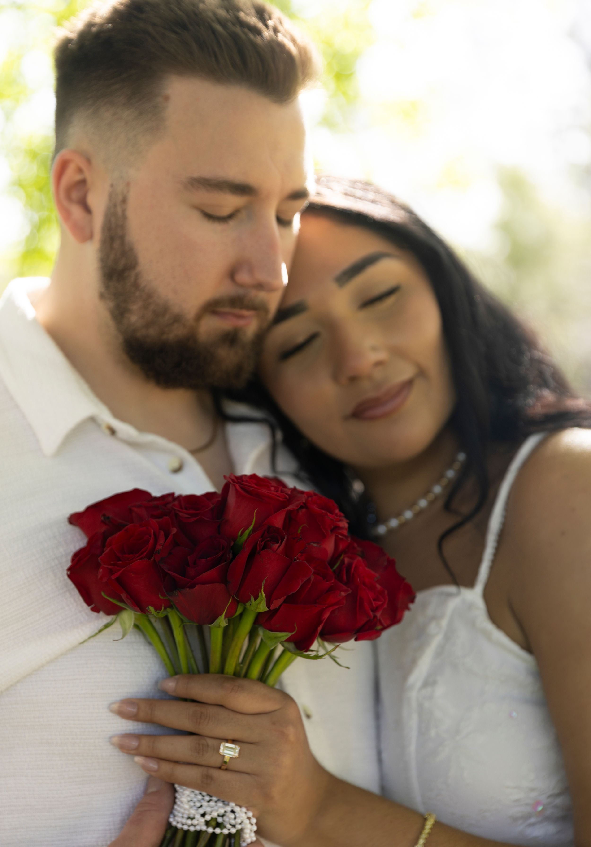 A romantic couple share an intimate moment while holding a bouquet of deep red roses in soft natural lighting.