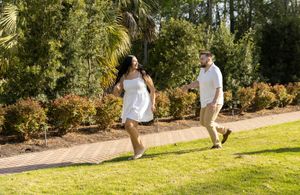 Two people walk together on a grassy path surrounded by lush tropical plants and palm trees in a garden setting.