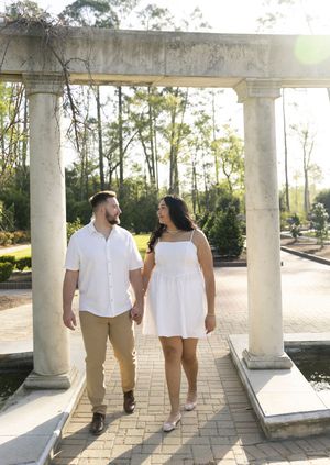 A couple wearing coordinating white outfits walks together under a columned pergola during golden hour.