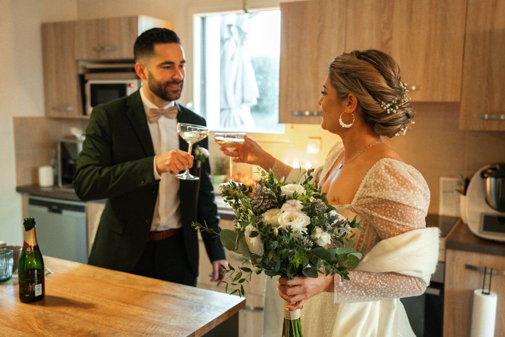 Wedding couple toasting with champagne glasses in modern kitchen with floral bouquet.