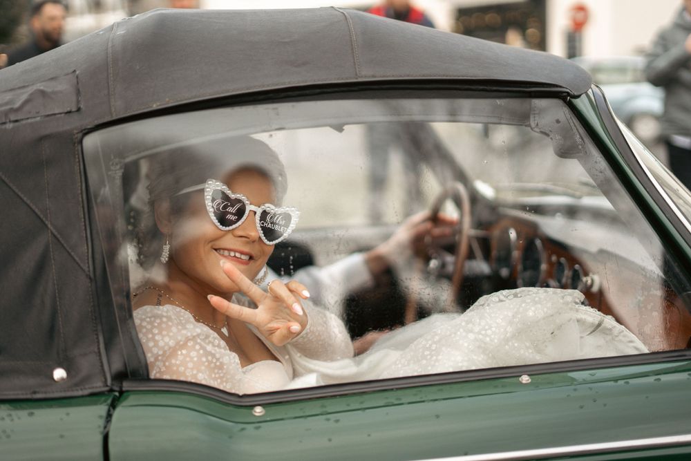 A stylish person in sunglasses and wedding dress sits in a vintage convertible car.