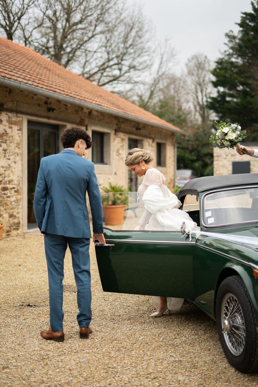A couple in wedding attire approaching a classic vintage green car outside a rustic stone building.