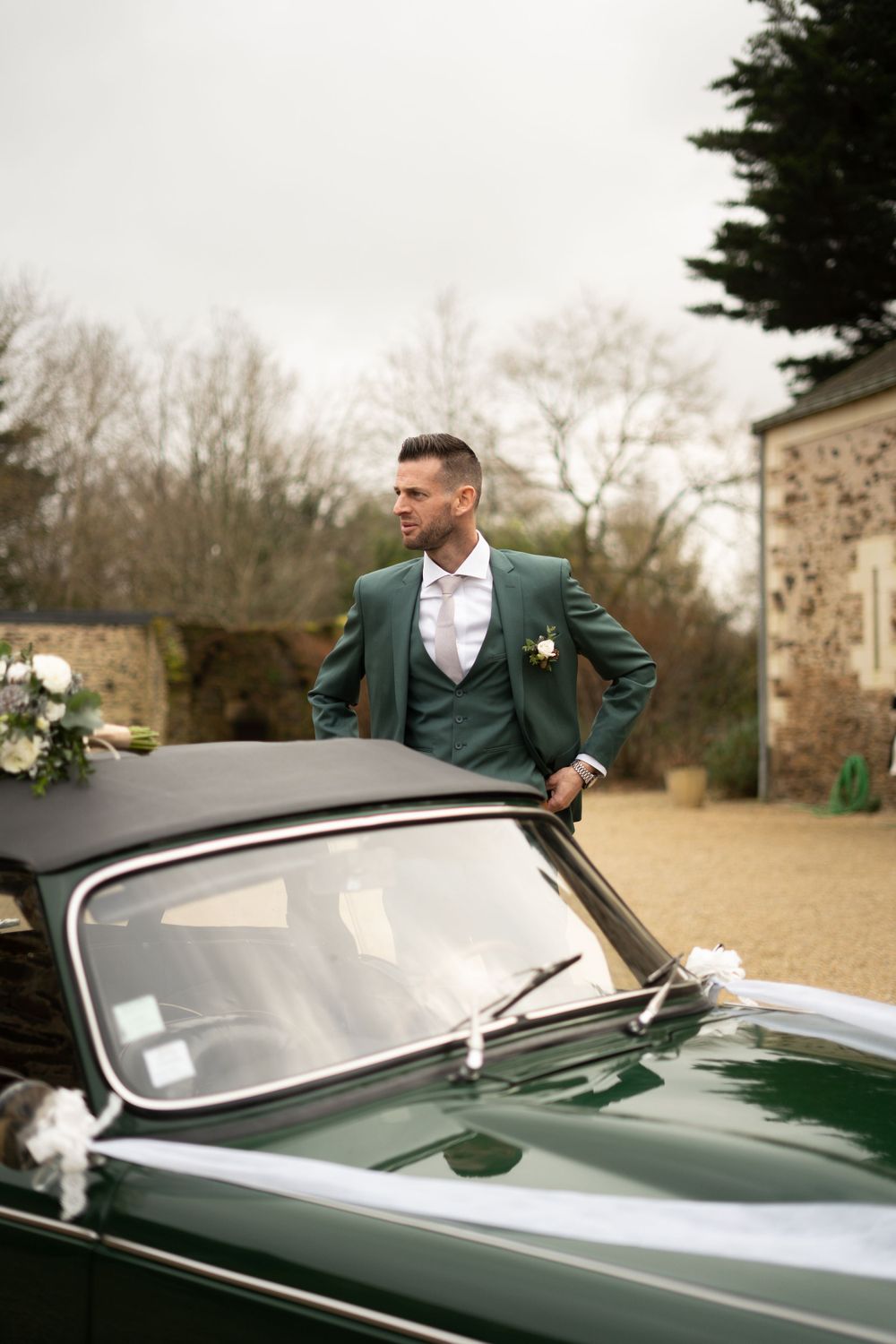 A classic green vintage car with wedding decorations parked outside a stone building.