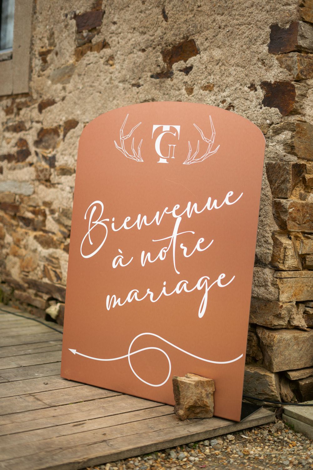 A terracotta-colored welcome sign with antler motif reads 'Bienvenue a notre mariage' against stone wall.