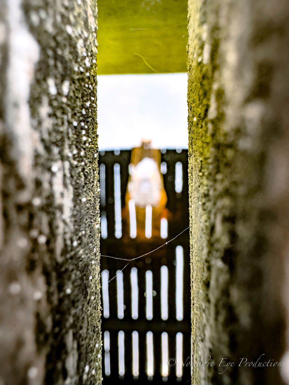 View through a narrow concrete gap showing a sunlit gate in the distance.