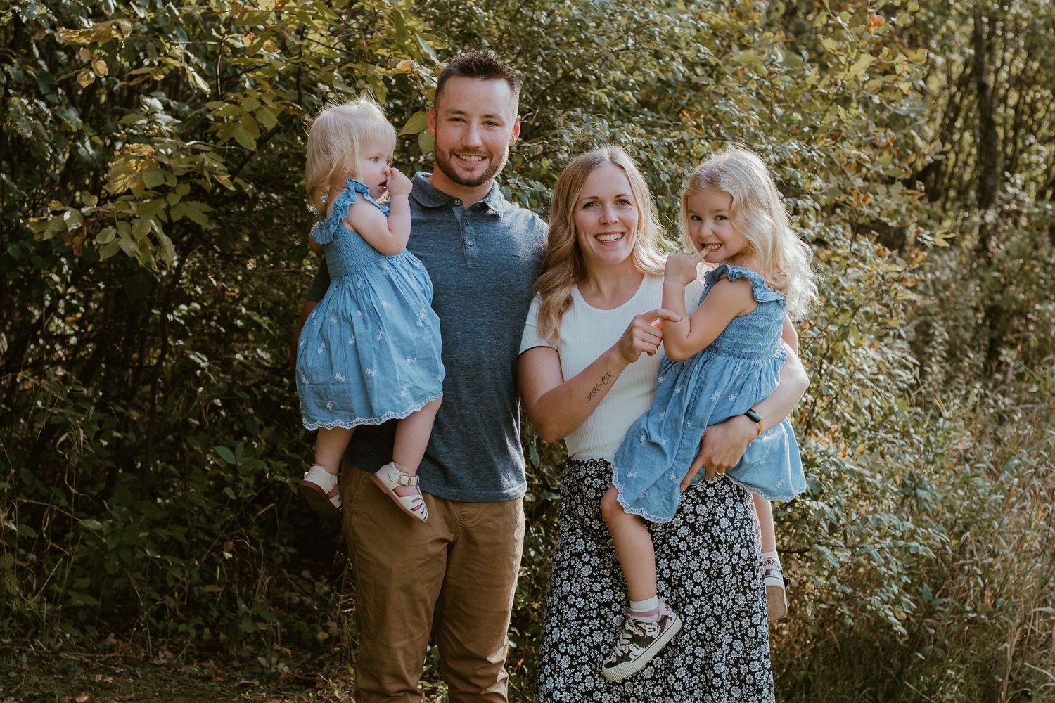 A family poses together outdoors in a natural setting with greenery and trees in the background.