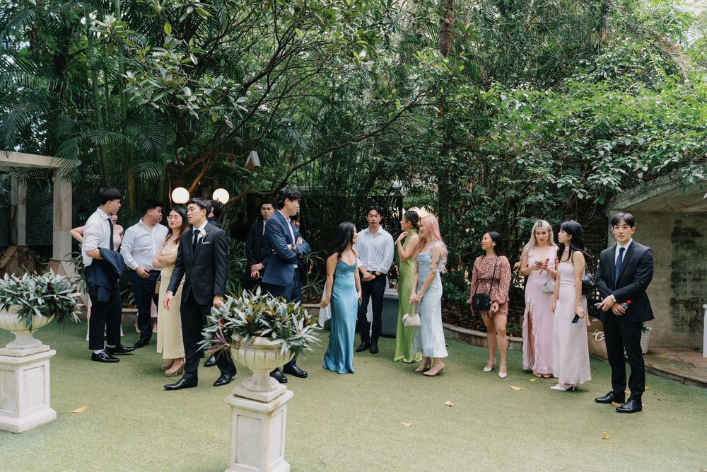 Wedding guests gather in a lush garden setting wearing formal attire and mingling during an outdoor reception.