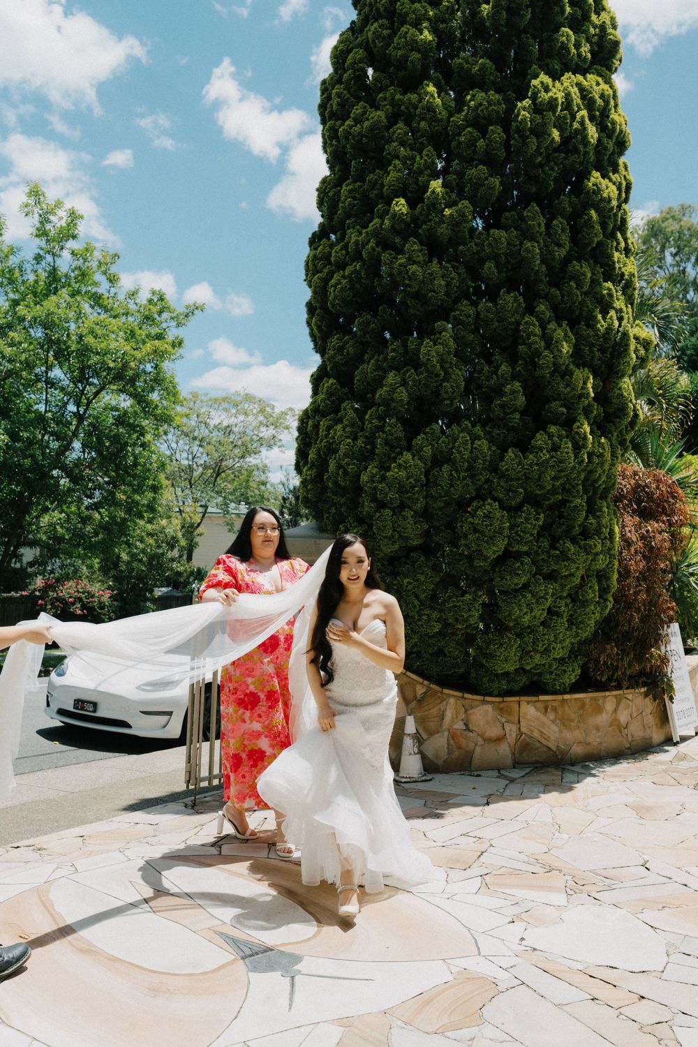 Two people in formal attire walk on a stone patio near a tall cypress tree and parked Tesla on a sunny day.