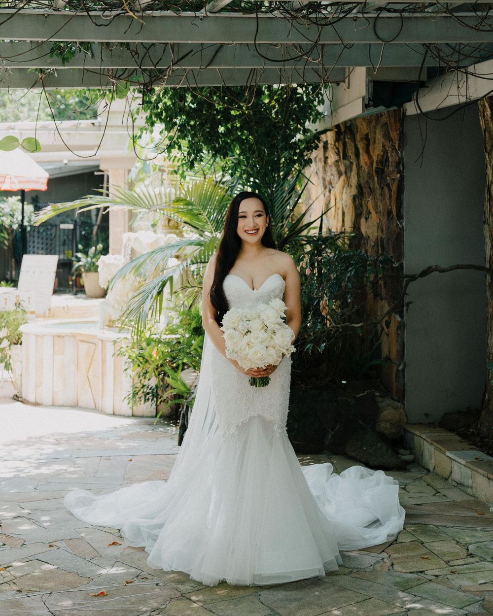 A bride in a white mermaid-style wedding gown with delicate lace details stands on a stone patio surrounded by greenery.