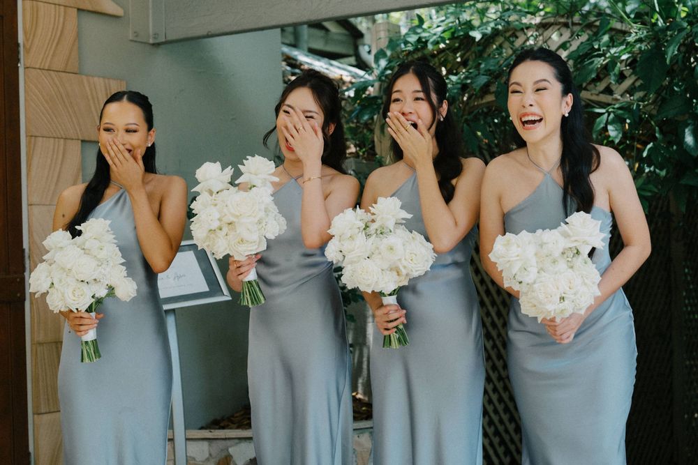 Four bridesmaids in light gray dresses hold white bouquets while laughing together during outdoor wedding photos.