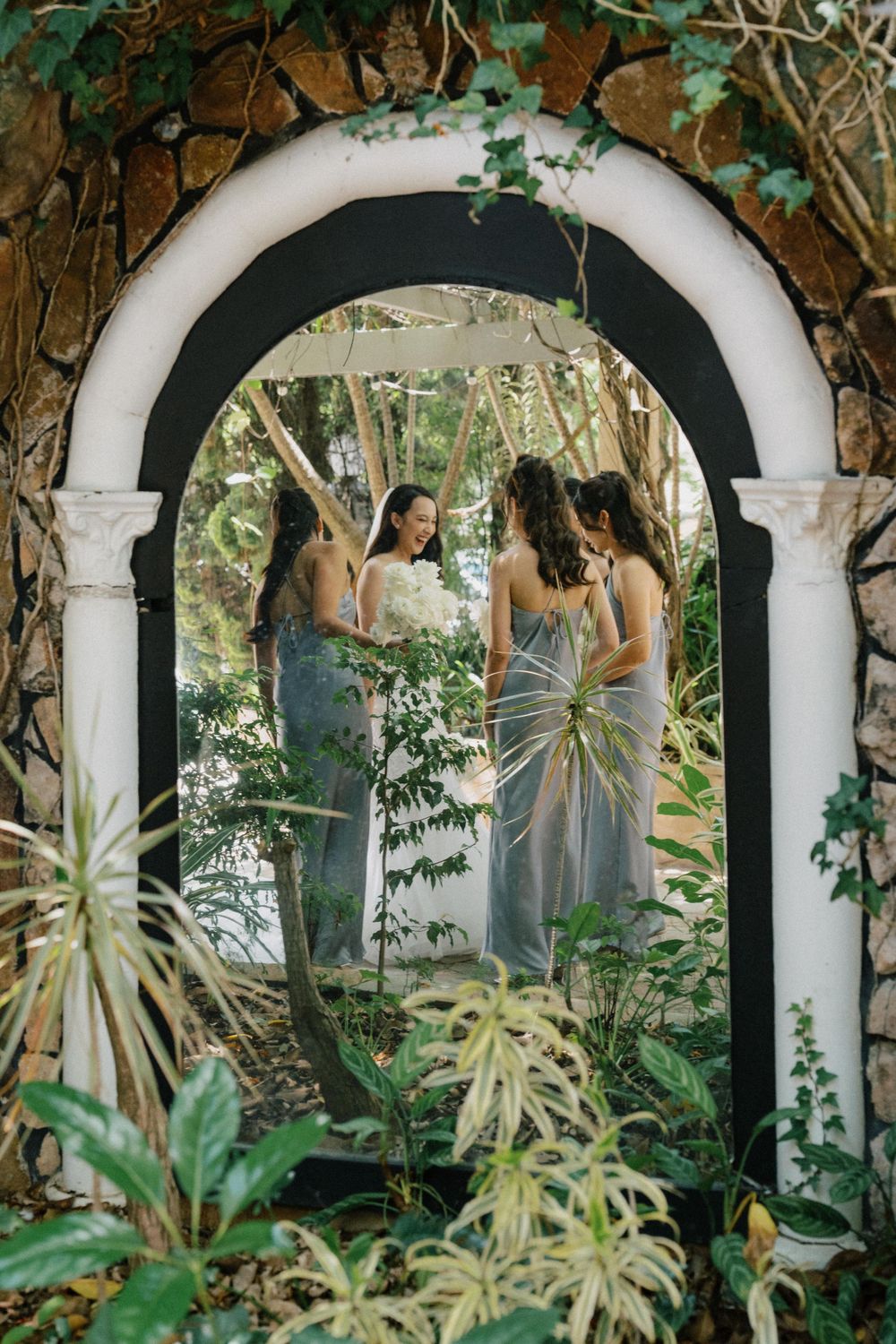 An elegant white arch with green vines frames people in flowy dresses standing in a lush garden setting.