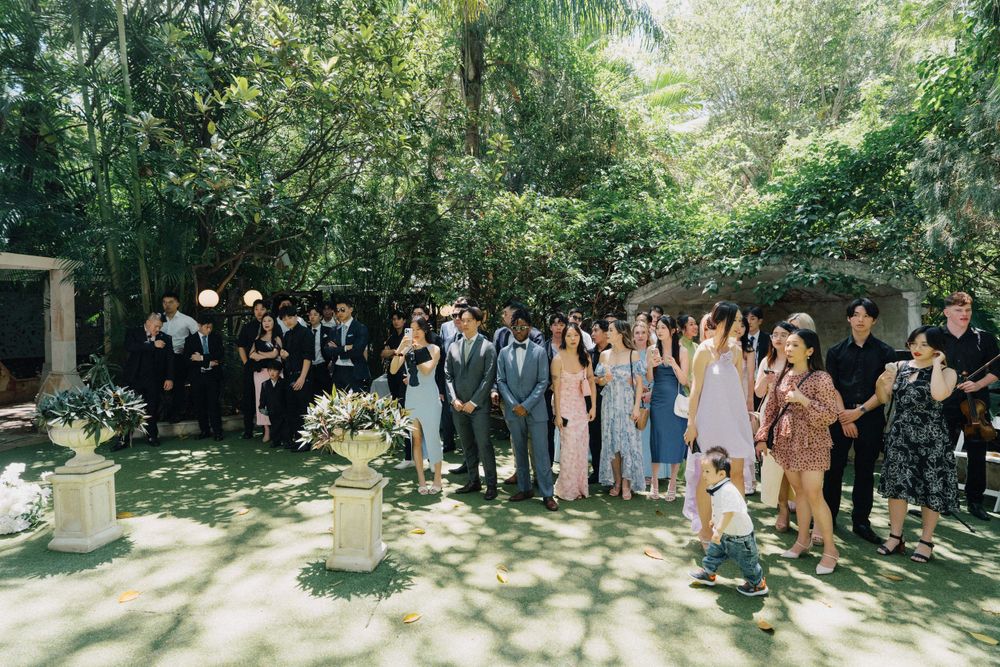 Large group of wedding guests gathered outdoors for a photo beneath shady trees with white floral arrangements.