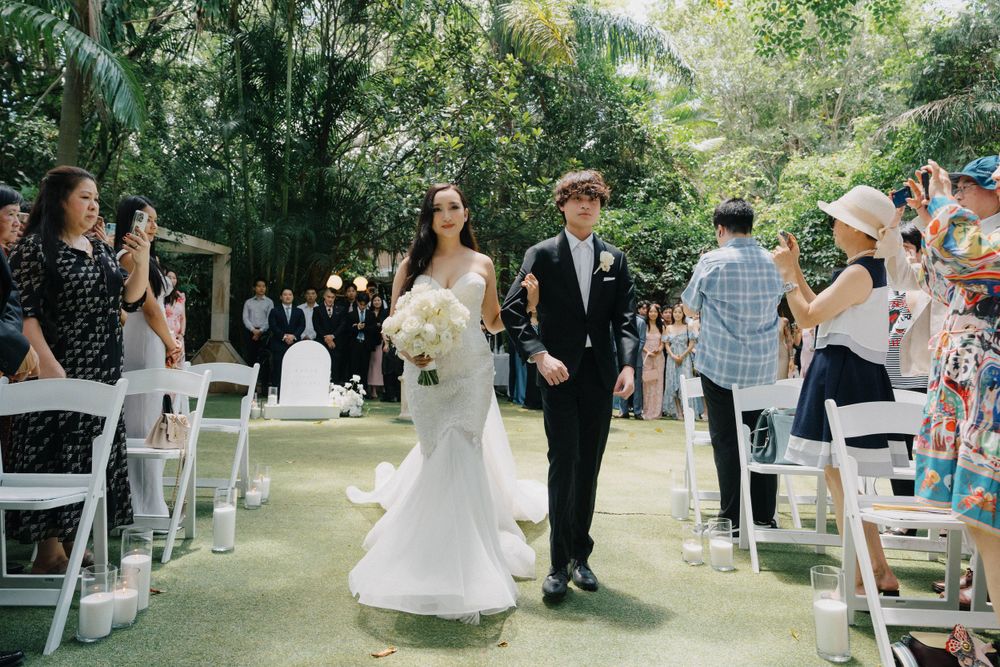 A wedding couple walks down an outdoor aisle lined with white chairs and tropical greenery at a garden ceremony.