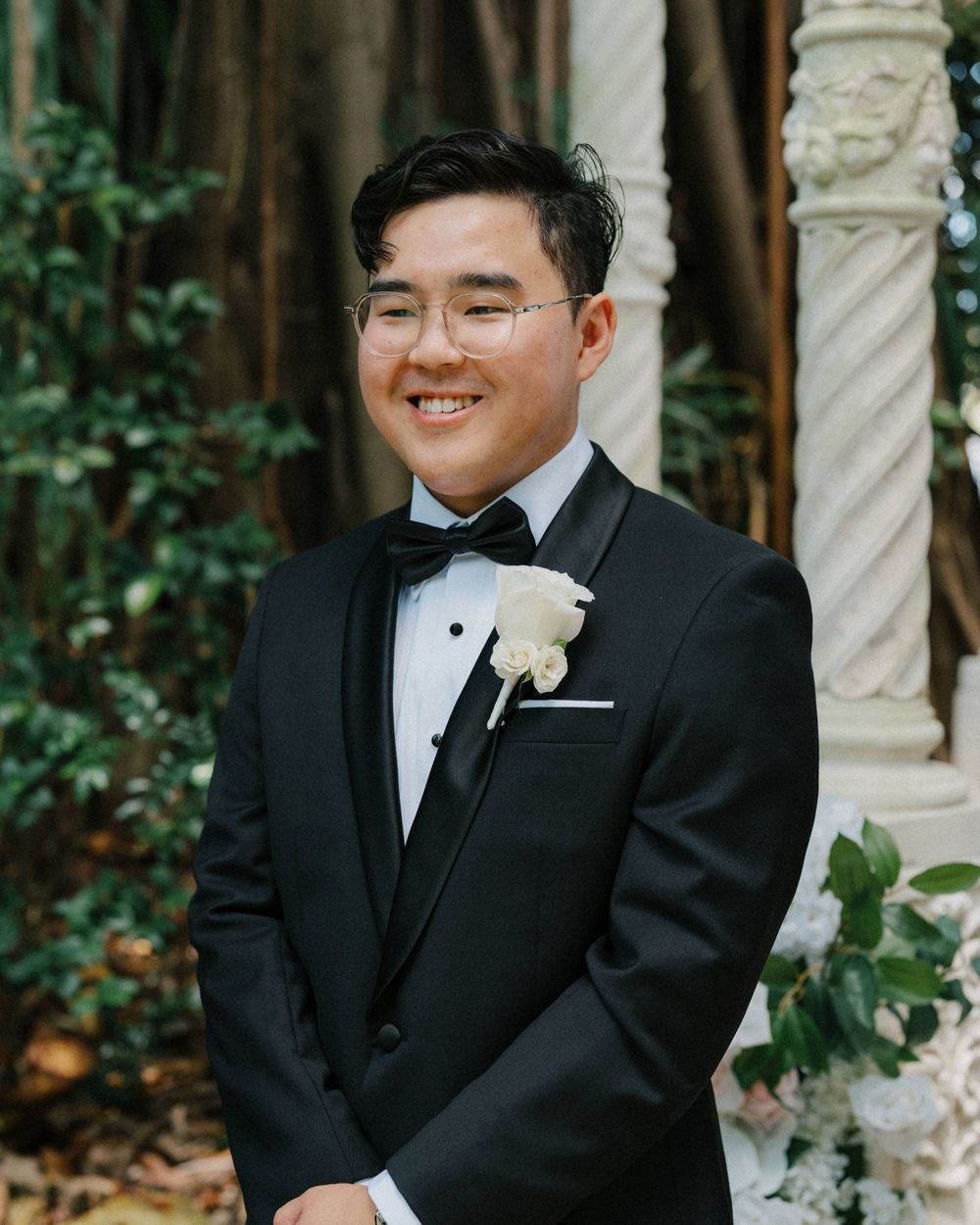 A person in a black tuxedo with glasses and white boutonniere smiling in front of greenery and stone columns.