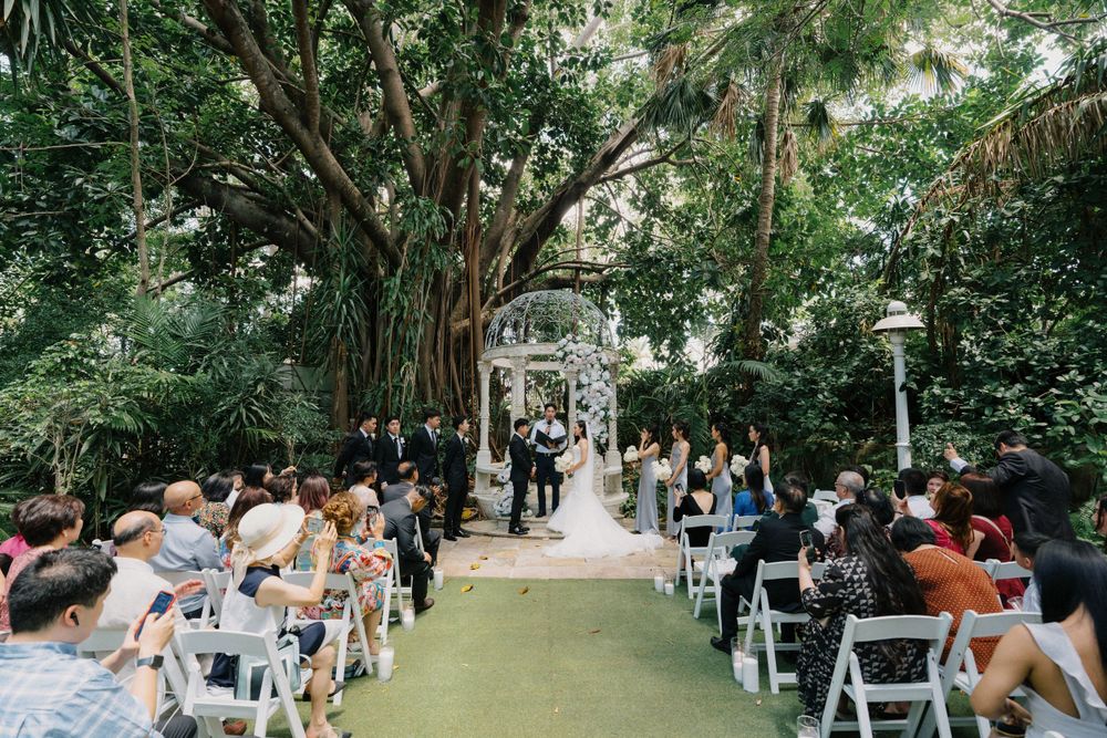 Outdoor wedding ceremony setup with white chairs under lush green trees and a winding stone pathway.