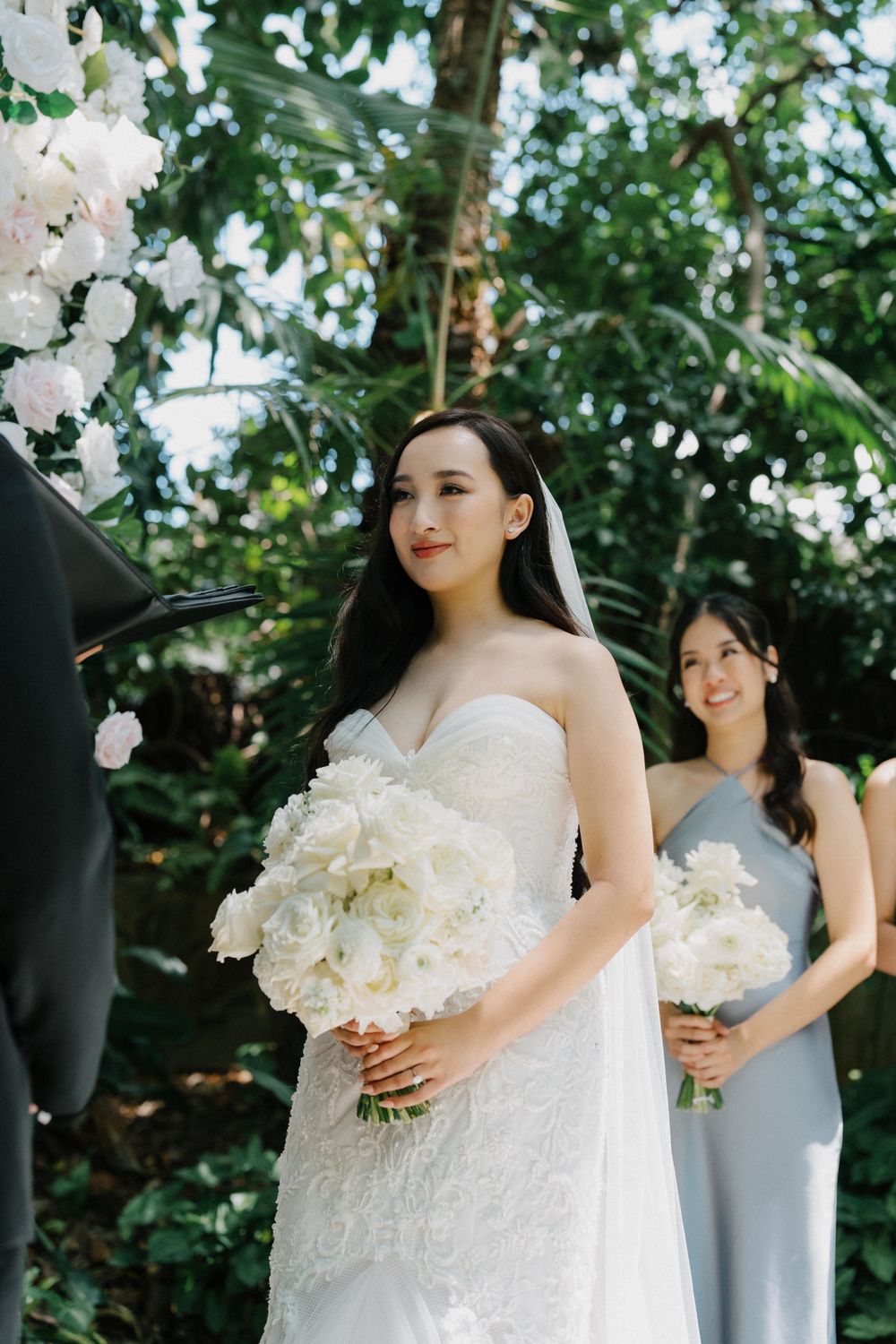 Outdoor wedding ceremony with white floral arrangements surrounded by lush greenery and trees.